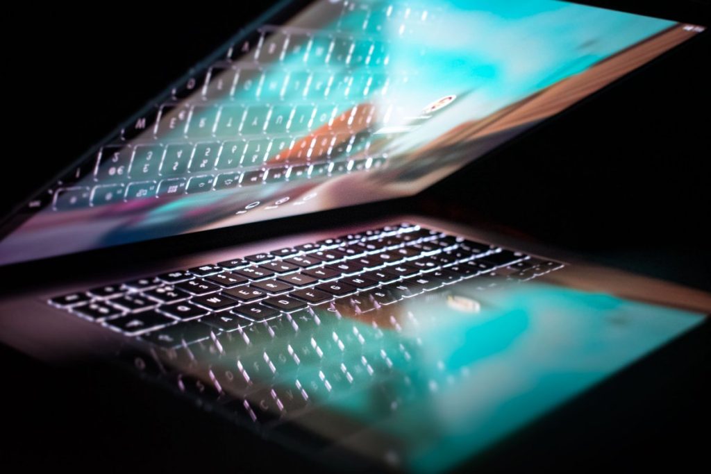 A partially closed laptop with illuminated keys reflects a bright screen against a dark background.