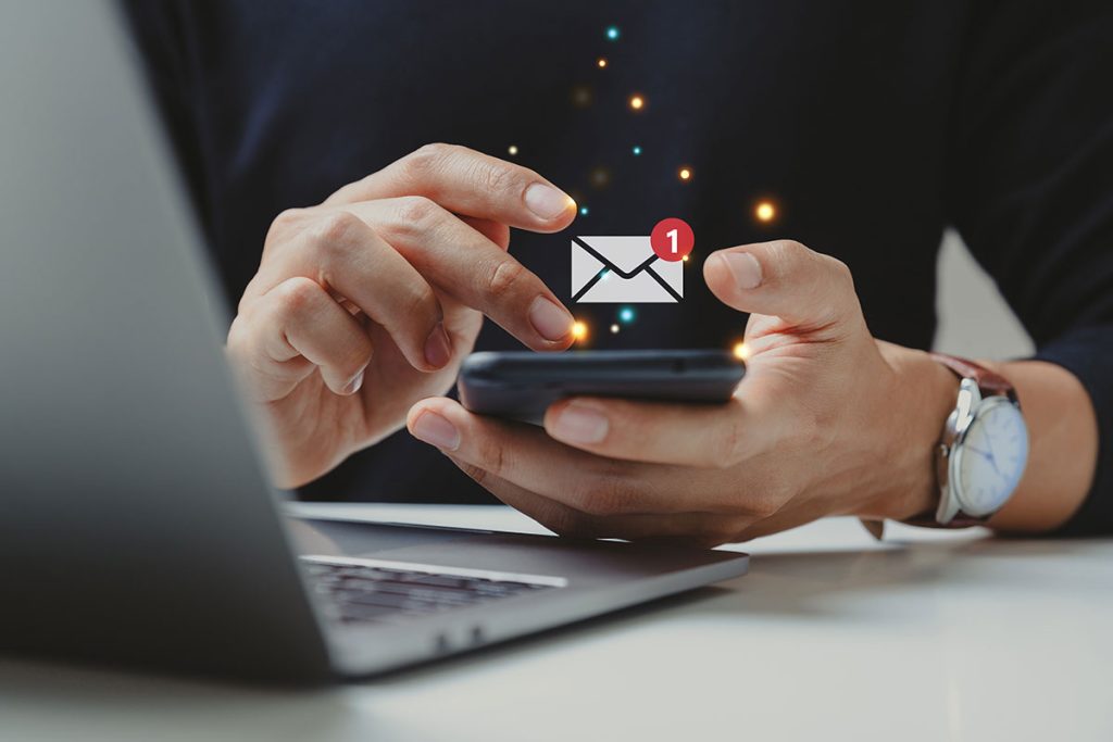 A person sits at a desk with a laptop, holding a smartphone that displays a new email notification icon, akin to receiving an exclusive concierge SMS.