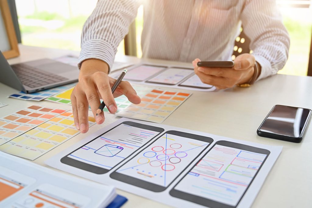A person is reviewing mobile app wireframes on paper, holding a smartphone and pointing at design layouts on a desk filled with color swatches and a laptop, seamlessly integrating elements of website design into the creative process.